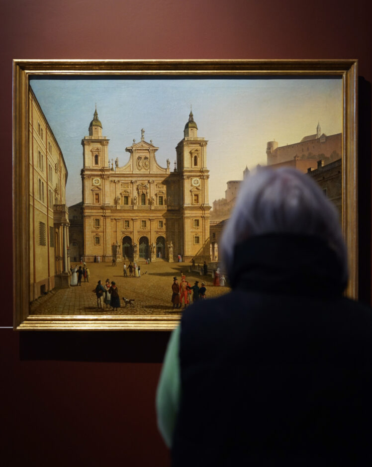 Eine Person betrachtet das Ölgemälde Der Domplatz mit Mariensäule und Domfassade in Salzburg von Johann Michael Sattler.