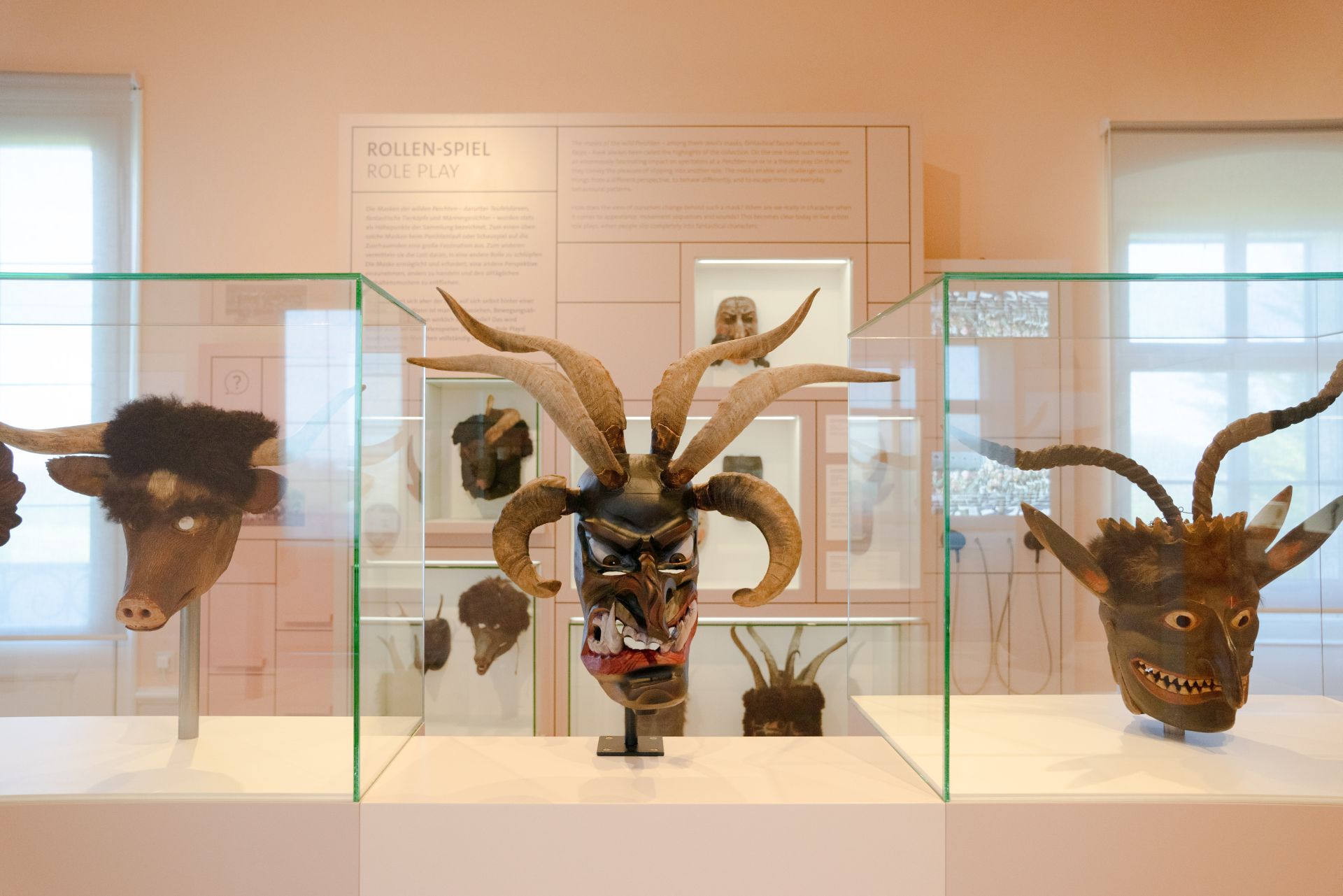 Drei gefürchtete und kunstvoll geschnitzte Masken mit Hörnern in Glasvitrinen vor einer Wand mit der Aufschrift „ROLLEN-SPIEL / ROLE PLAY“.