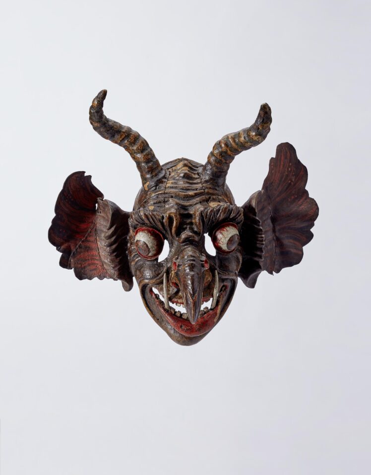 „Teufels“-Maske, mit zwei geschnitzten Widderhörnern, Lederohren;  die bunte Fassung teilweise von dunkler Patina überdeckt; Sehschlitze neben den Glotzaugen; dünnwandig.

Gehörnte Maske mit großen abstehenden Lederohren. Dominante Geiernase mit zwei krebsartigen Geschwülsten an den Nasenflügeln und offenen Nasenlöchern, über der Nasenwurzel dekorativ gewulstete Falte, die sich mit den stark plastisch geriefelten Augenbrauen verbindet; weit heraustretende Glotzaugen. Die zu einem breiten Lachen verzogenen Lippen des großen Mundes entblößen zwei aus dem Unterkiefer wachsende lange Vampirzähne inmitten der regelmäßig angeordneten Zahnreihen oben (10 Zähne) und unten (8 Zähne); dicke, wulstige Unterlippe. Stark ausgebildete Wangen-, Augen- und Stirnfalten. Große abstehende Lederohren: an die geschnitzten Ohrmuscheln sind mächtige Drachenohren aus Leder angefügt.  
Die Innenseite der extrem dünnwandigen Maske ist fast zur Gänze mit aufgeklebten Leinenstreifen ausgekleidet.
Erwerb: 1858 Schenkung als erste Maske in die Sammlung des Städtischen Museum Carolino Augusteum