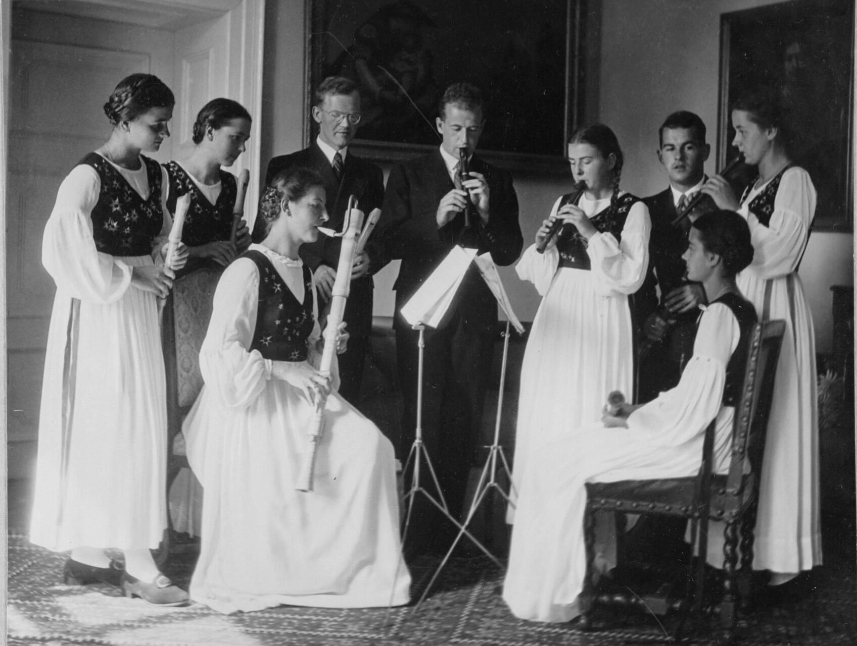 Historisches Schwarz-Weiß-Foto einer Gruppe von jungen Frauen und Männern in traditioneller Kleidung, die Blockflöten spielen.