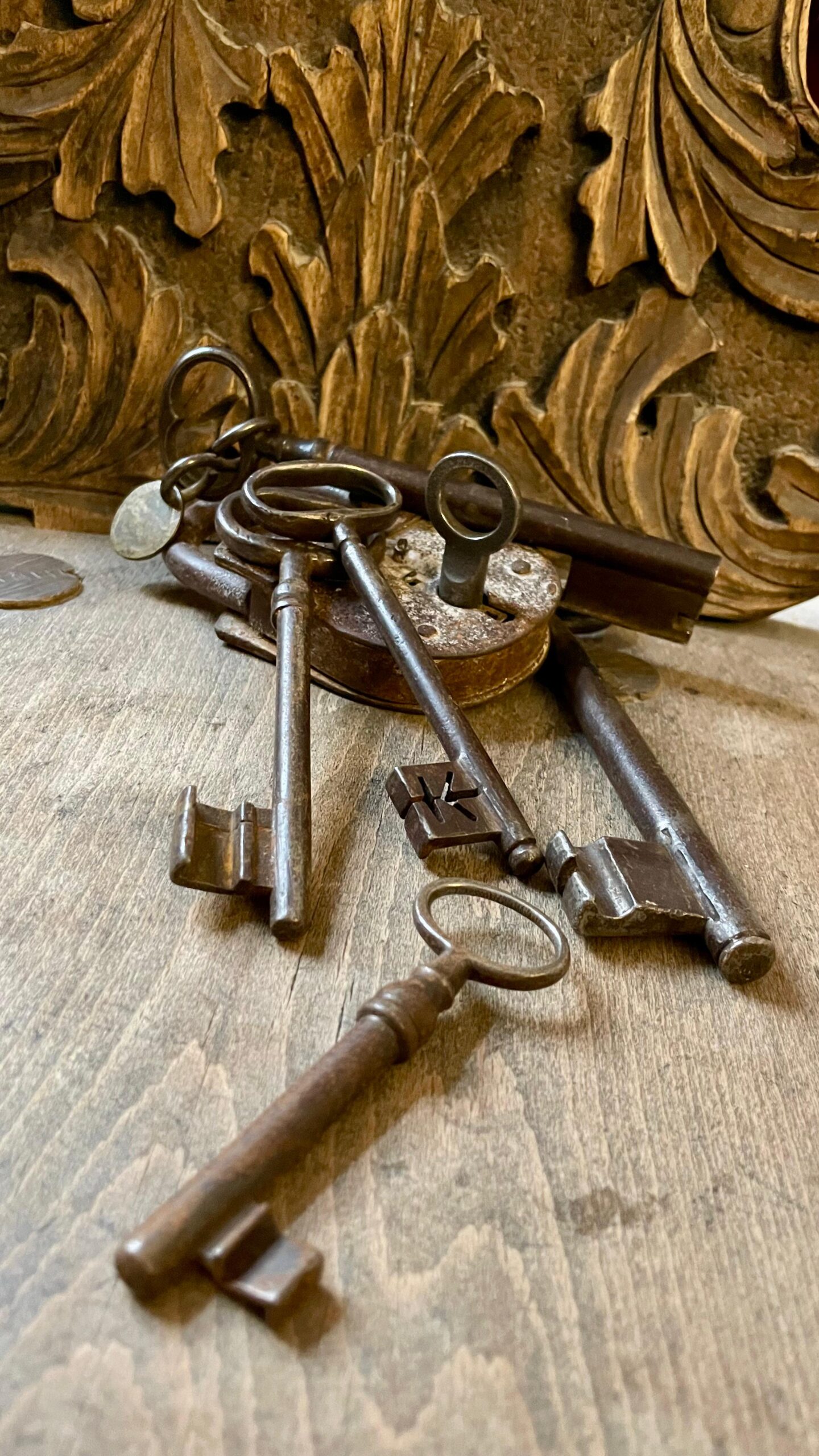 Mehrere antike, rostige Schlüssel und ein altes Vorhängeschloss liegen auf einer hölzernen Oberfläche vor einem geschnitzten Ornament.