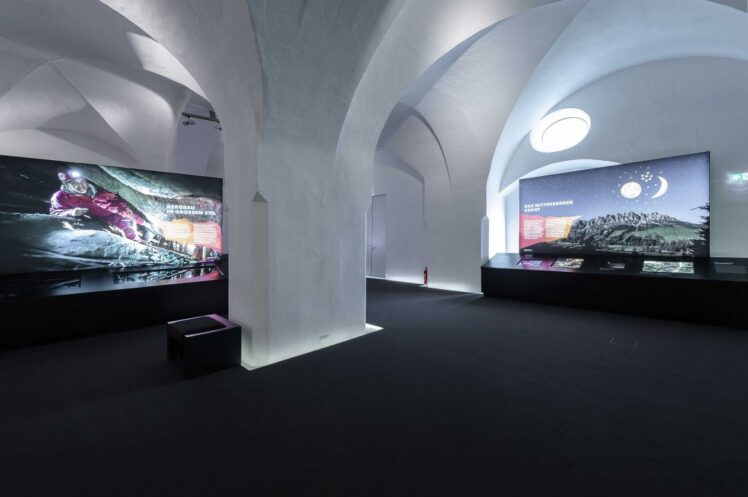 Blick durch einen historischen Gewölbesaal der Museumsausstellung mit beleuchteten Medientafeln und dunklem Teppichboden