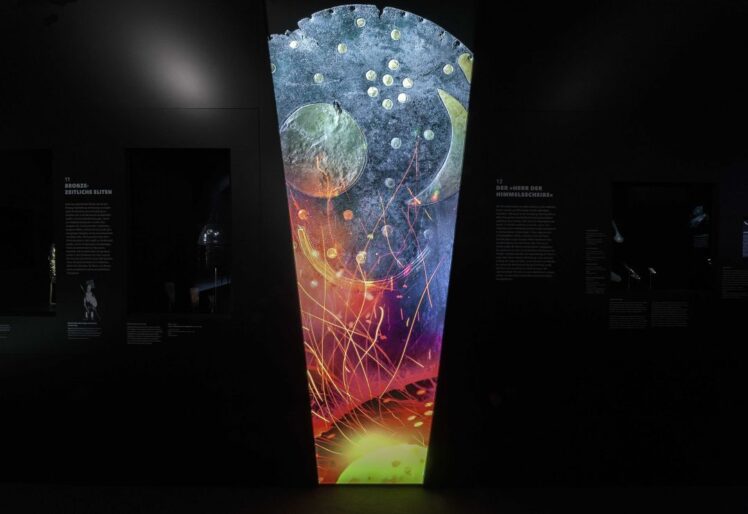 Beleuchtete vertikale Vitrine oder Projektionswand mit der Himmelsscheibe von Nebra und bunten Lichteffekten in einer dunklen Museumsausstellung