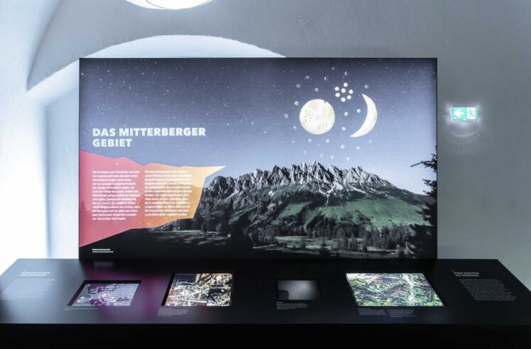 Große Ausstellungstafel mit der Aufschrift "Das Mitterberger Gebiet" vor einer alpinen Bergkulisse bei Nacht und integrierten Medientischen