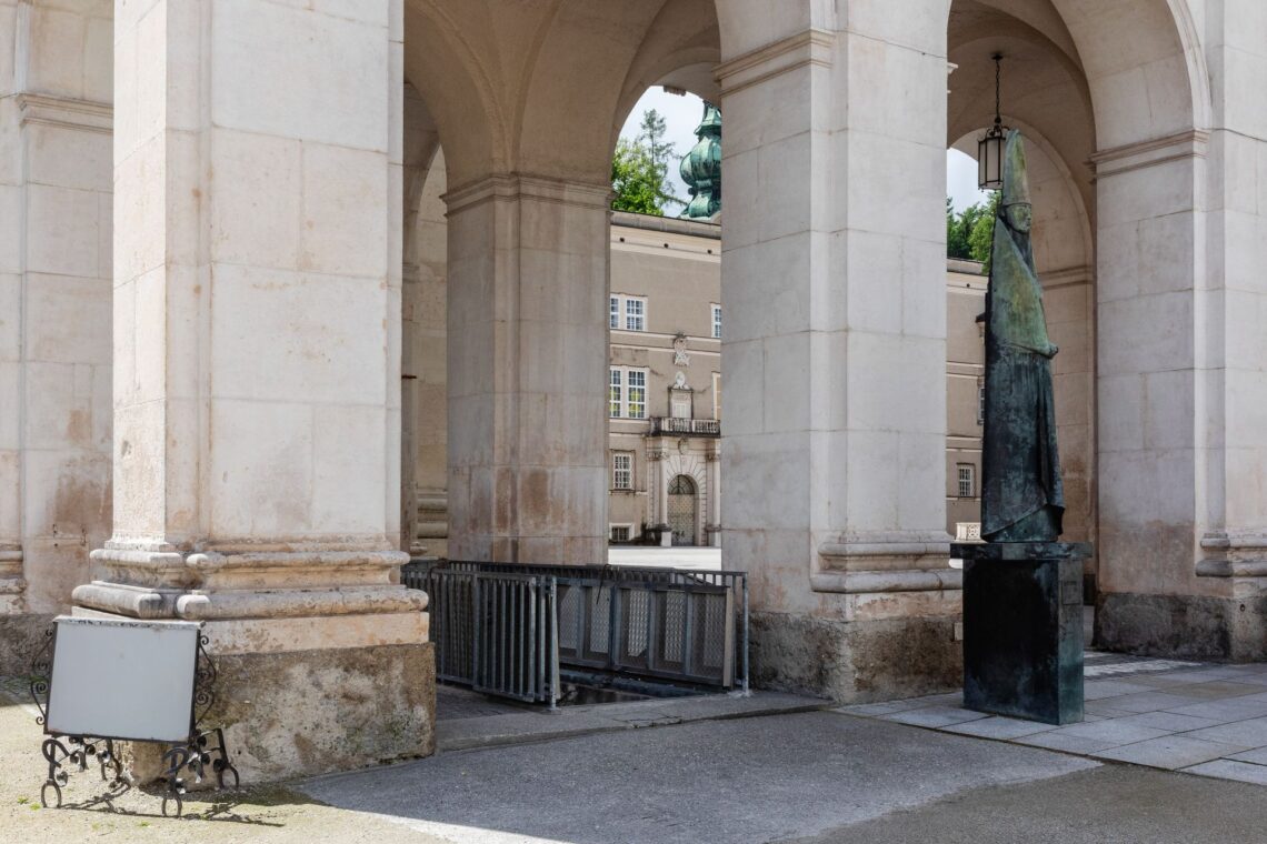 Blick durch massive Arkadensäulen auf einen Platz mit einer dunklen Skulptur und historischen Gebäuden im Hintergrund