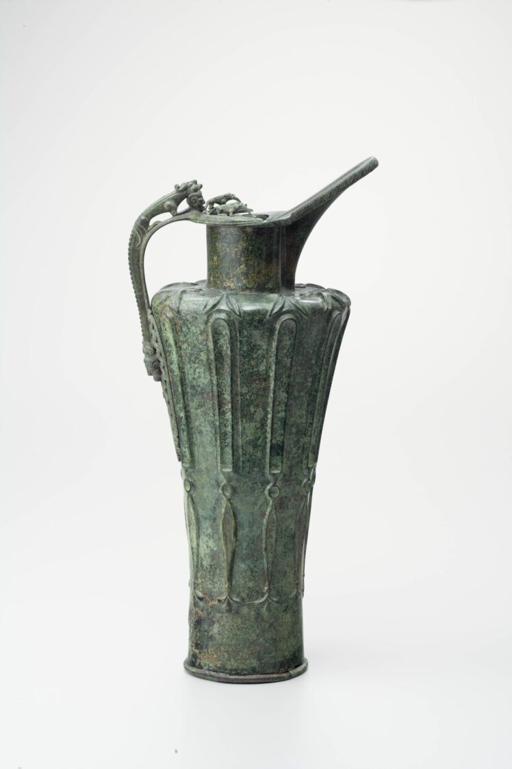 A slender, historical bronze pitcher with a long spout, vertical fluting, and a figural handle with a lid against a white background.