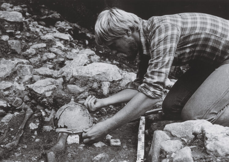 Black-and-white photograph of Kurt Zeller kneeling at an archaeological excavation site, carefully uncovering a heavily weathered helmet from the soil.