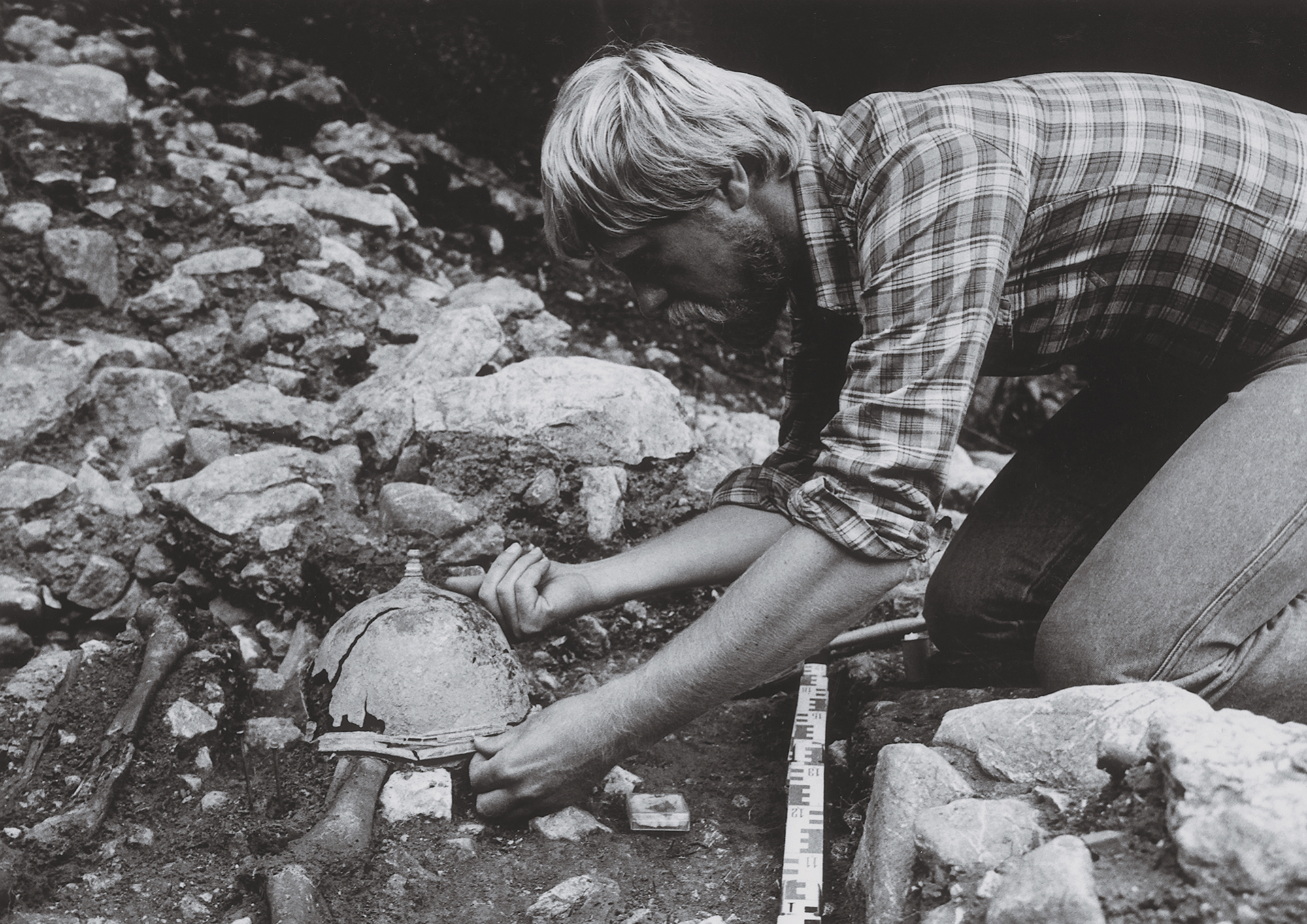 Black-and-white photograph of Kurt Zeller kneeling at an archaeological excavation site, carefully uncovering a heavily weathered helmet from the soil.