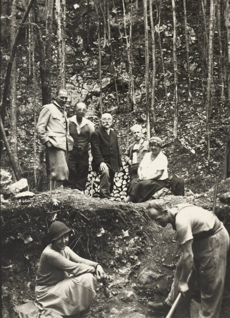 Historical black-and-white photograph of a group of seven people at an archaeological excavation site in a wooded area, with two individuals working in the soil in the foreground.