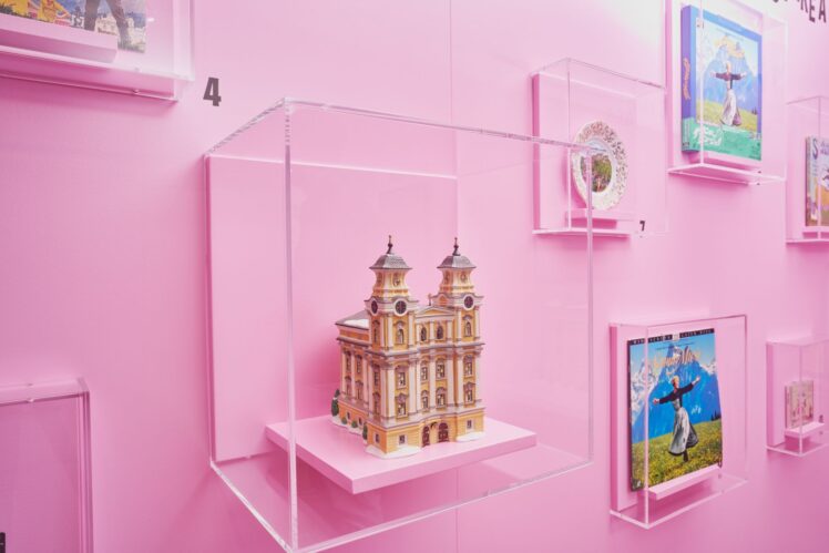 Rosafarbener Ausstellungsbereich mit einem Architekturmodell, Sammelteller und internationalen „The Sound of Music“-Veröffentlichungen in Vitrinen.