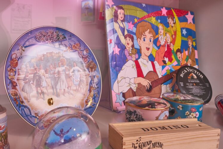 Vitrine mit internationalen „The Sound of Music“-Fanartikeln, darunter Sammelteller, Schallplatten, Dosen und eine farbenfrohe Animationsdarstellung.