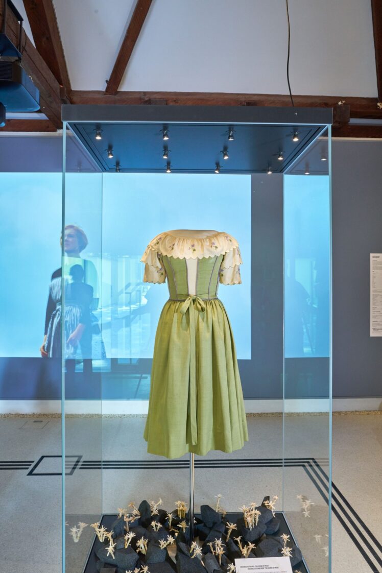 Grünes Filmkostüm aus „The Sound of Music“ in einer Glasvitrine, mit einer Filmszene im Hintergrund.