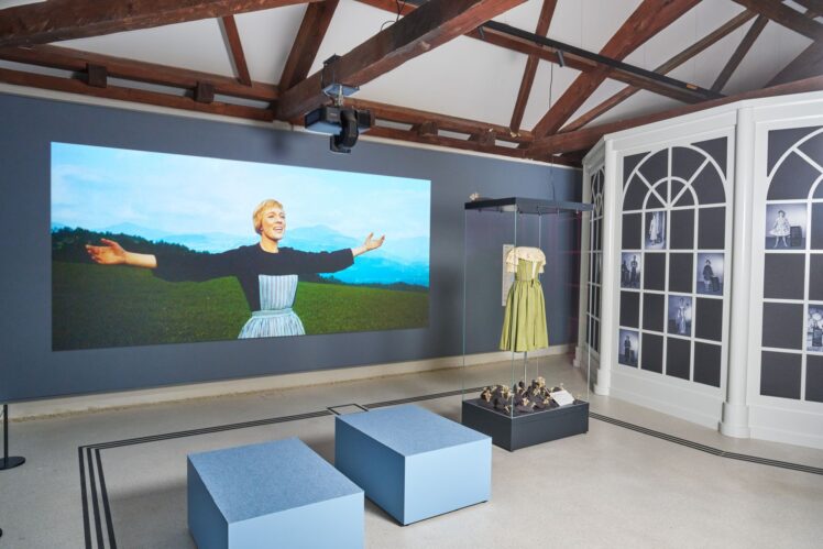 Ausstellungsraum zu „The Sound of Music“ mit großformatiger Filmprojektion, einem Kostüm in einer Glasvitrine und historischen Fotografien.