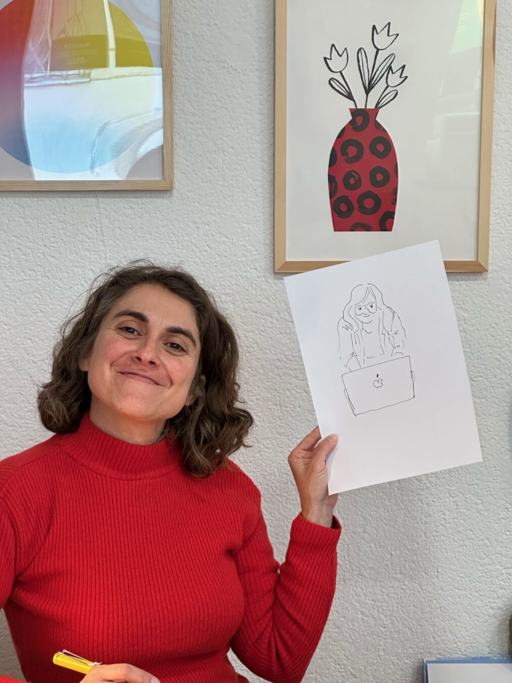 Gemma Palacio lächelt in die Kamera und hält eine Zeichnung hoch, die eine Person hinter einem Laptop zeigt. Im Hintergrund hängen gerahmte Illustrationen an der Wand.