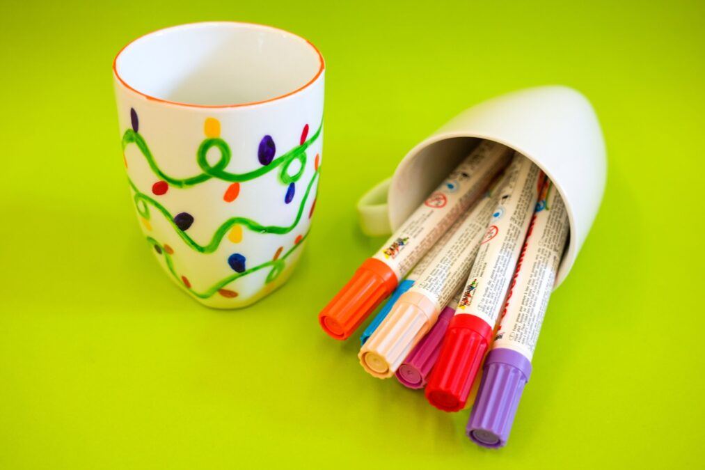 Handbemalte weiße Tasse mit buntem Lichterketten-Muster und eine umgekippte Tasse gefüllt mit Porzellanmalstiften auf grünem Hintergrund.