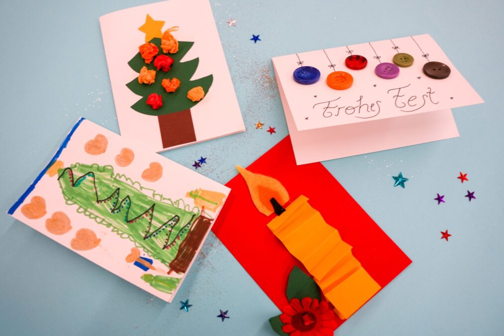 Ein Set aus vier handgemachten Weihnachtskarten, darunter eine mit einem Weihnachtsbaum aus Papierknäueln, eine mit Knopf-„Kugeln“ und der Aufschrift „Frohes Fest“, eine bemalte Kinderzeichnung und eine Karte mit einer 3D-Papierkerze, alles verstreut auf einem hellblauen Untergrund mit Konfetti.