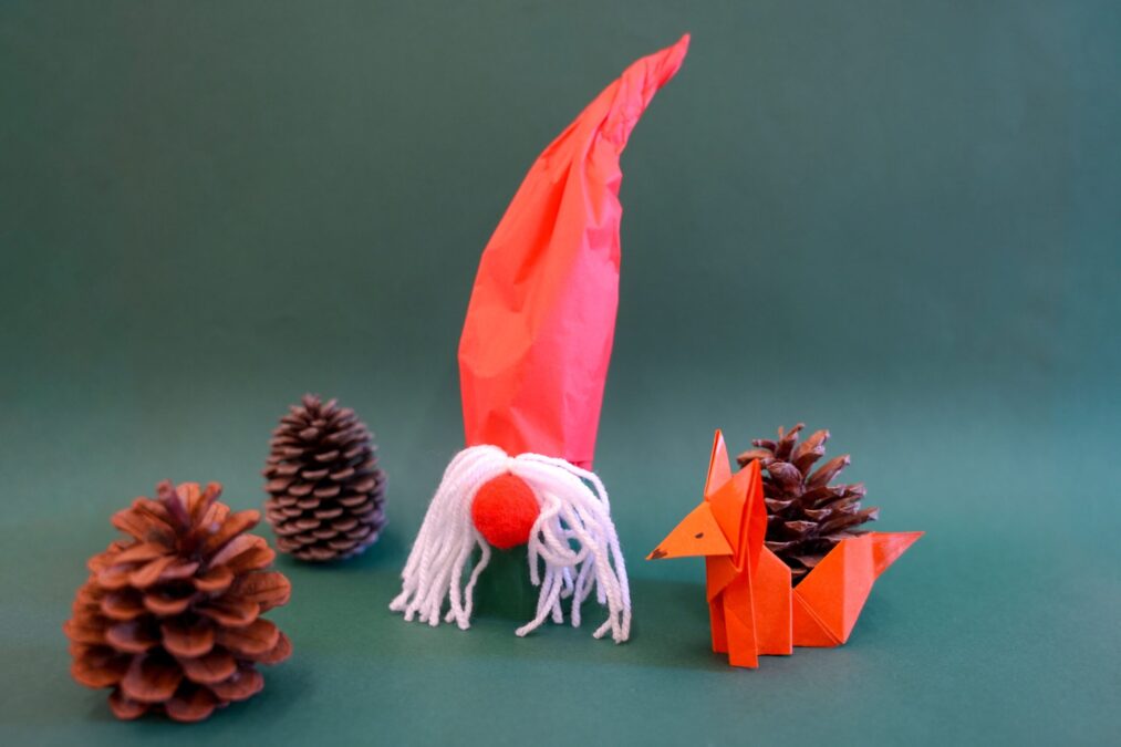 Ein selbstgebastelter Zwerg mit roter Mütze und weißem Wollbart, begleitet von einem Papier-Fuchs mit einem Tannenzapfen und weiteren Tannenzapfen auf grünem Hintergrund.