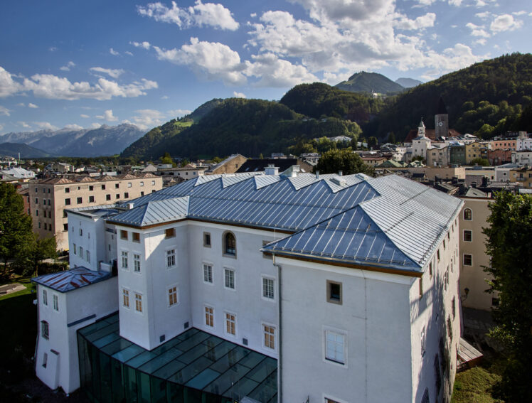 Blick auf das Keltenmuseum Hallein nach der Dachsanierung. Das historische Gebäude mit weißer Fassade und erneuertem silbernem Metalldach ist vor der Kulisse der Stadt Hallein und der umliegenden Berglandschaft zu sehen.