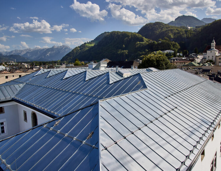 Blick auf das Keltenmuseum Hallein nach der Dachsanierung. Das historische Gebäude mit weißer Fassade und erneuertem silbernem Metalldach ist vor der Kulisse der Stadt Hallein und der umliegenden Berglandschaft zu sehen.
