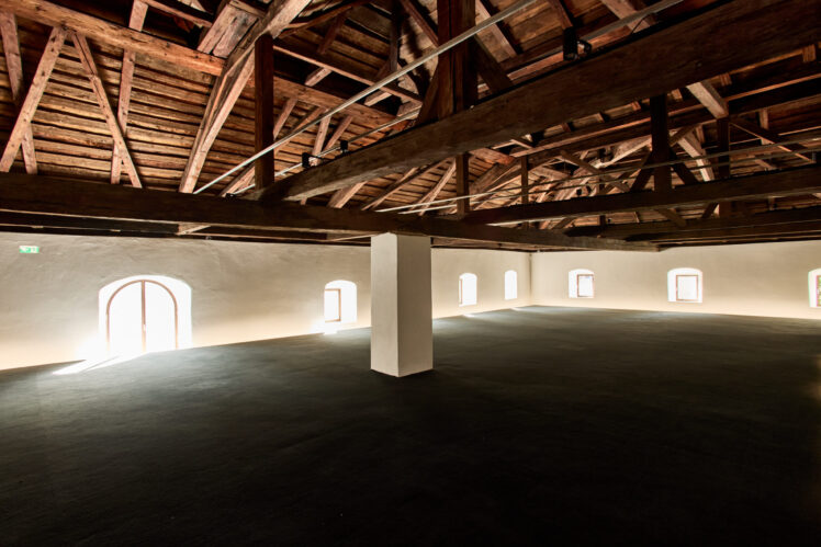 Großzügiger Veranstaltungs- und Ausstellungsraum im Kurt Zeller Forum des Keltenmuseums Hallein. Ein historischer Dachstuhl mit massiven Holzbalken überspannt den offenen Raum mit weißen Wänden, dunklem Boden und mehreren Rundbogenfenstern.