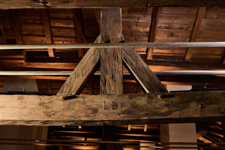 Detailansicht des historischen Dachstuhls im Kurt Zeller Forum des Keltenmuseums Hallein. Mehrere massive Holzbalken mit eingeritzten historischen Zeichen und Markierungen treffen aufeinander. Davor verlaufen horizontale Stahlseile.