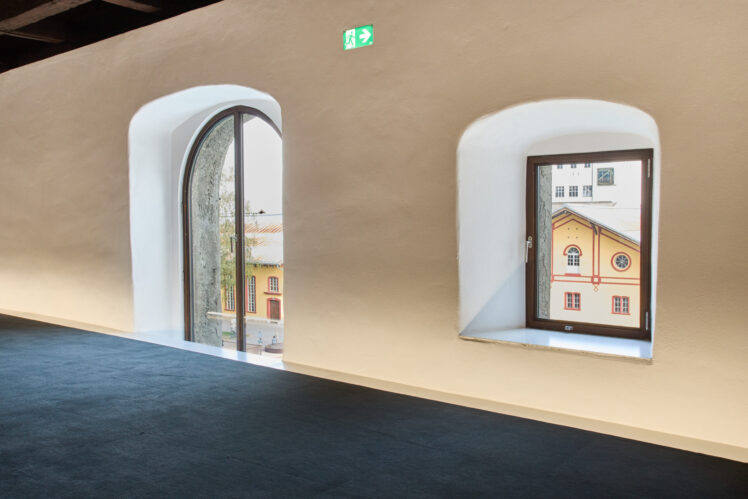 Blick aus dem Kurt Zeller Forum im Keltenmuseum Hallein auf die Pernerinsel. Durch ein hohes Rundbogenfenster und ein rechteckiges Fenster sind die gelben Fassaden mit roten Verzierungen der historischen Salinengebäude zu sehen.