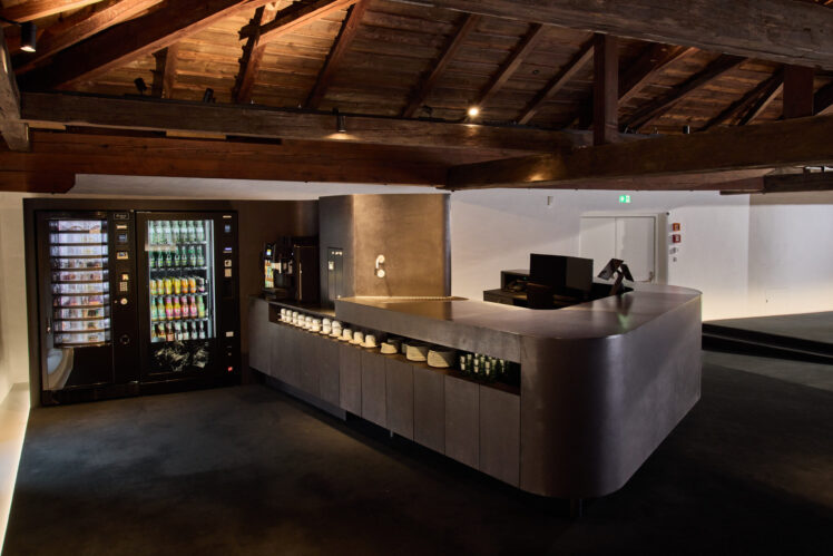 Blick in das Bistro des Kurt Zeller Forums im Keltenmuseum Hallein. Eine moderne, dunkle Theke mit Geschirr und Kaffeeausgabe steht unter einem historischen Dachstuhl mit massiven Holzbalken. Links befinden sich Getränkeautomaten.