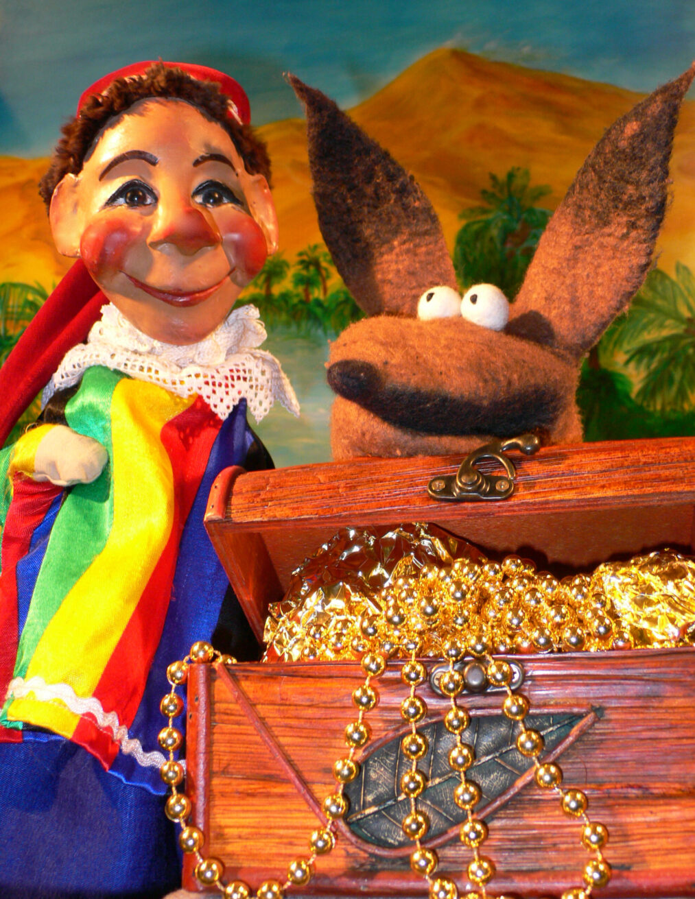 Eine lächelnde Handpuppe im Narrenkostüm und eine Stoffpuppe eines Fuchses mit großen Augen stehen hinter einer offenen Schatztruhe voller goldener Perlenketten und Folie vor einem gemalten Wüstenhintergrund.